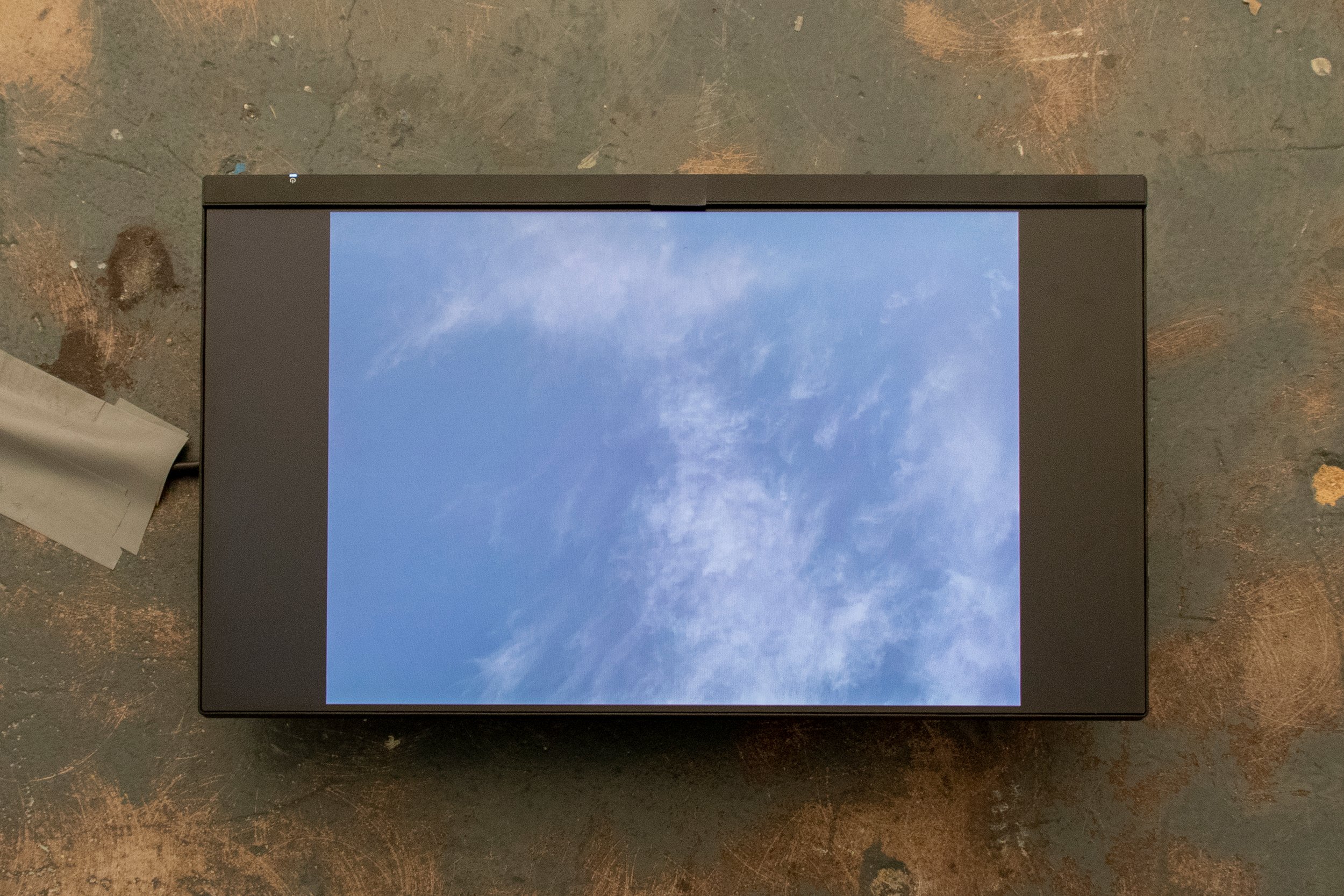 Small horizontal video monitor lying on a worn surface and displaying a pale blue sky with wispy clouds.