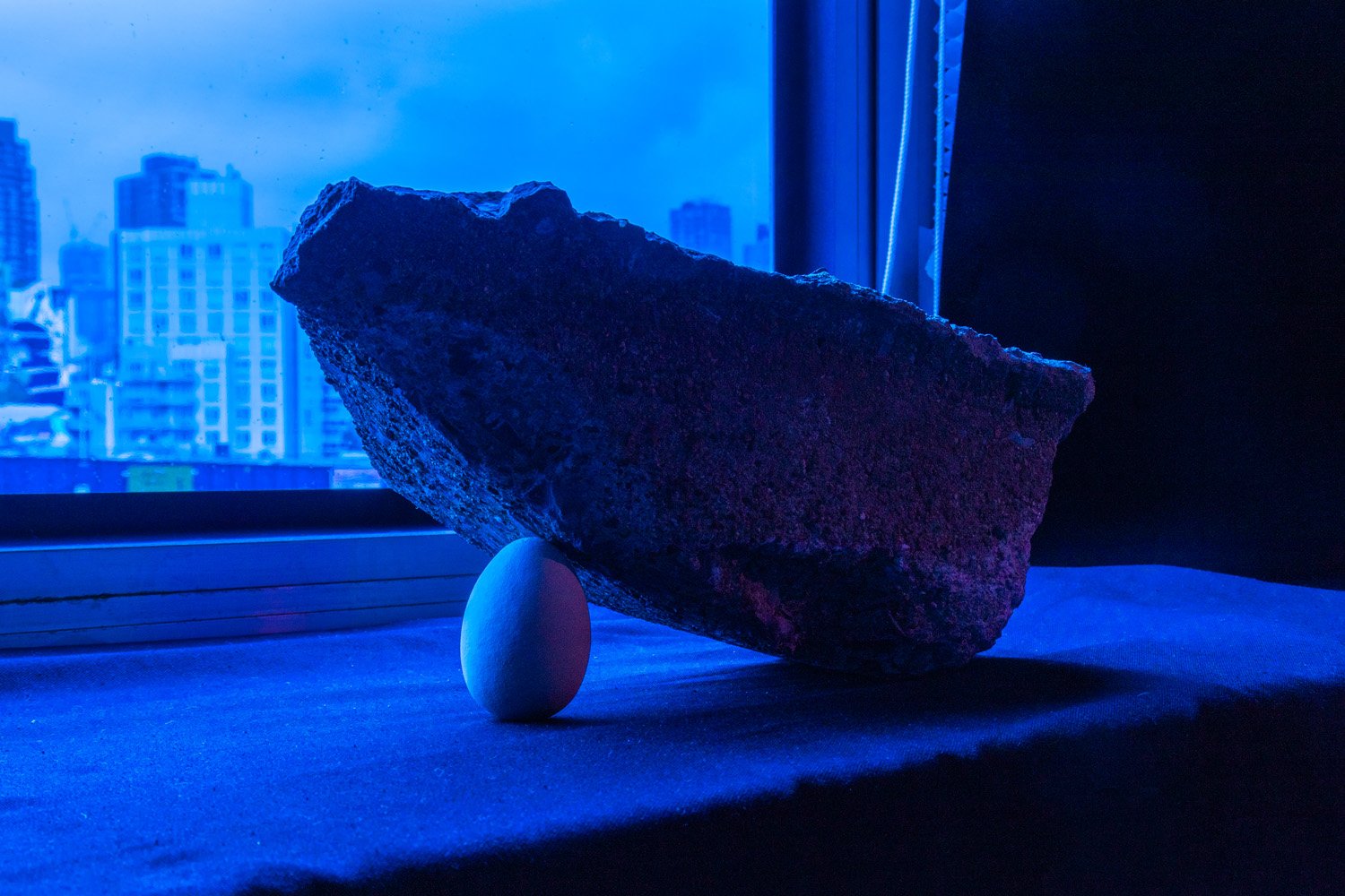 White egg supporting the edge of a large rough stone on a windowsill illuminated by deep blue light.
