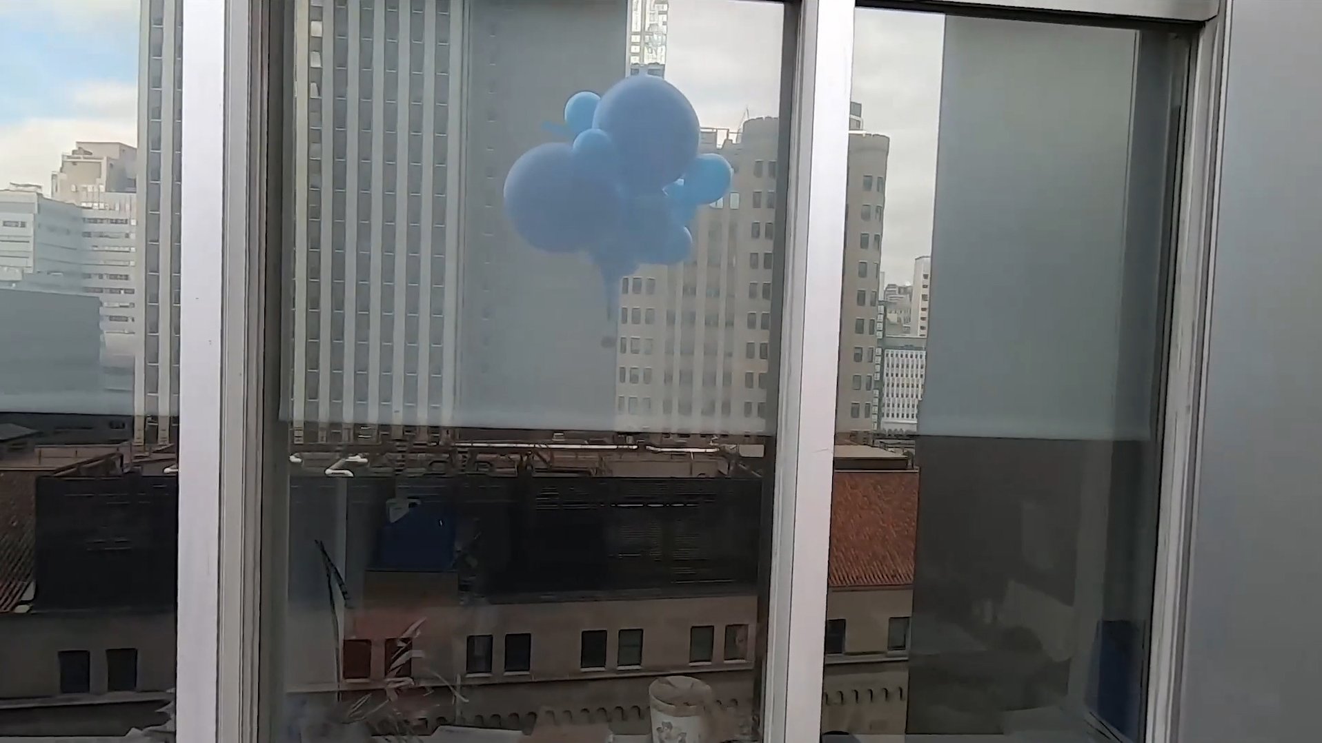 Video still showing a cluster of blue balloons floating outside a high-rise window with city buildings visible beyond.