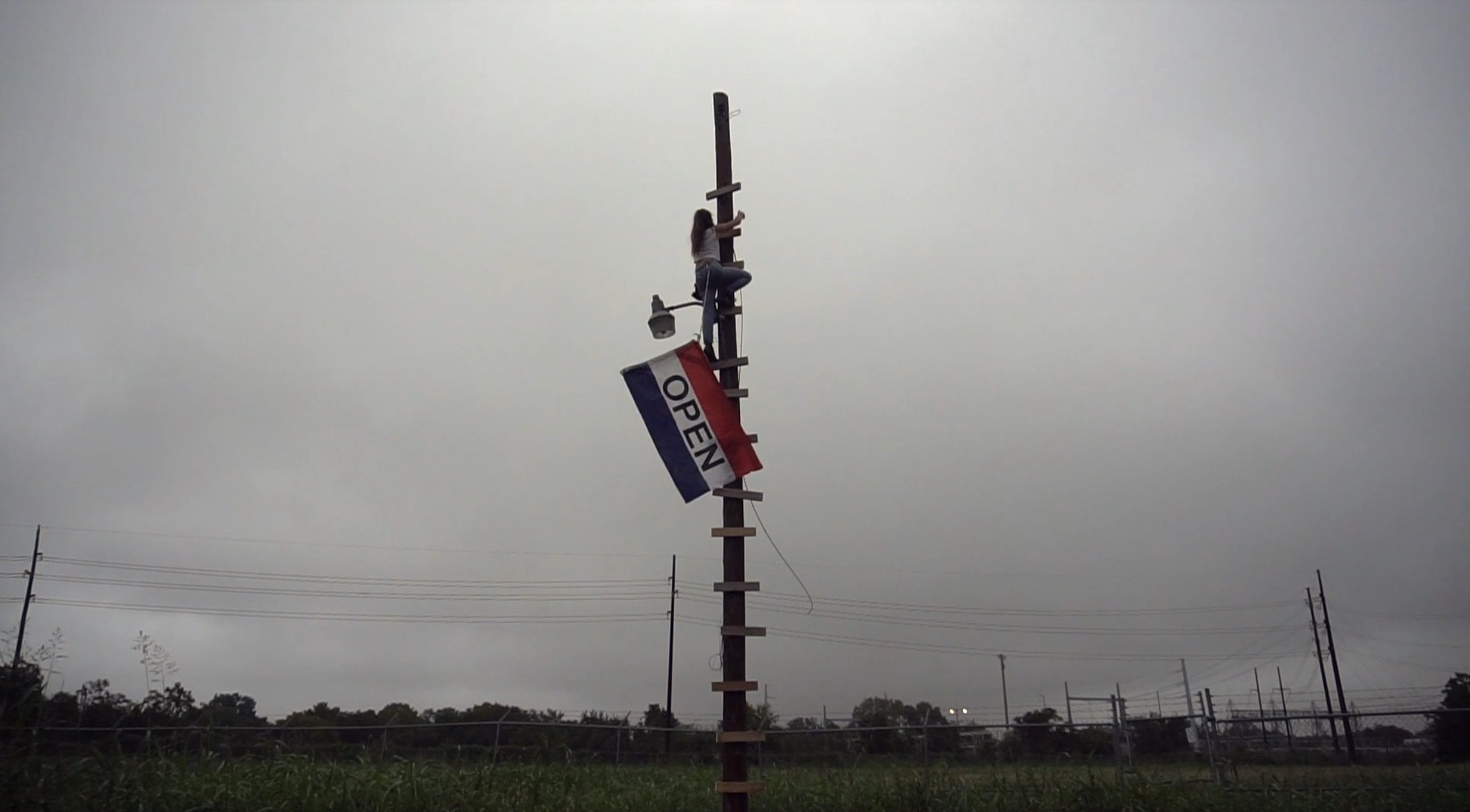 Performance still showing a person climbing a tall wooden pole in a grassy field beside a large red, white, and blue flag reading OPEN.