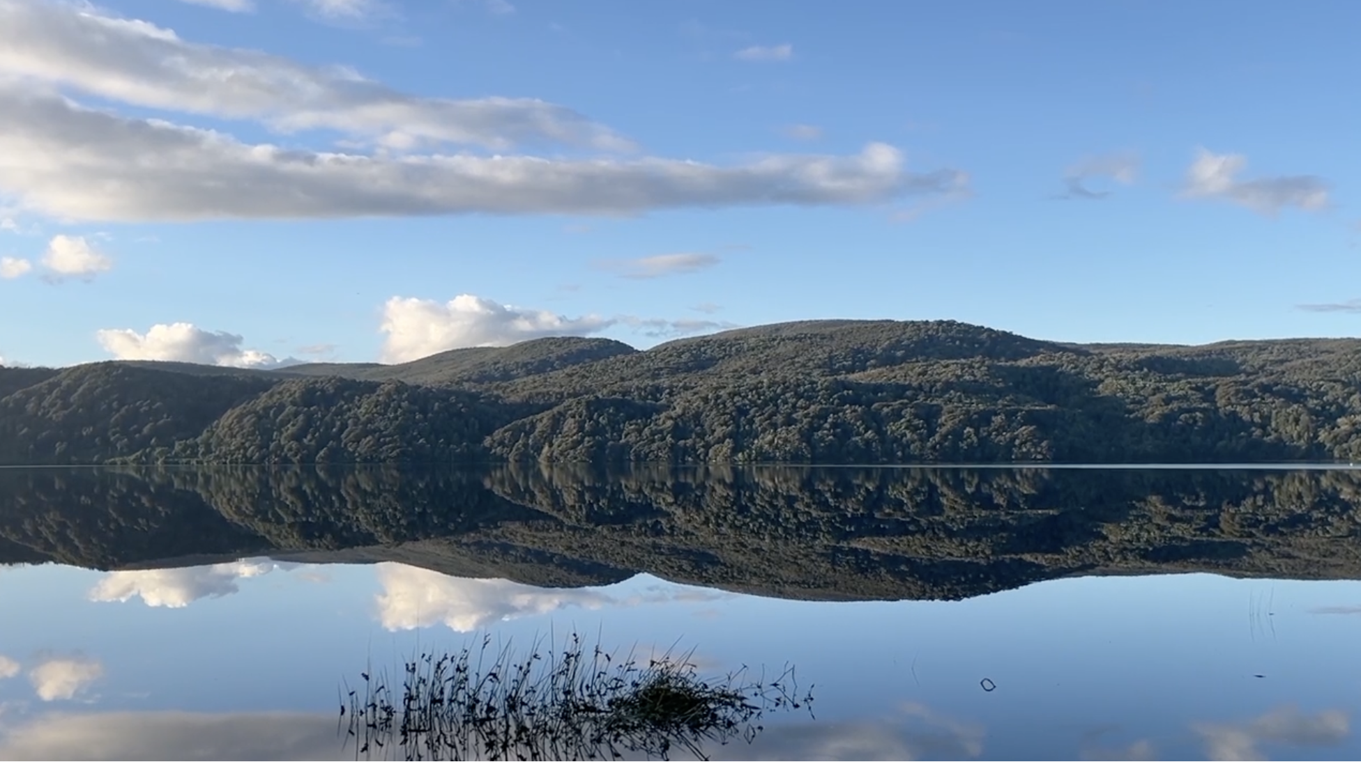 Still lake reflecting forested hills and a blue sky with clouds, creating an almost symmetrical landscape.