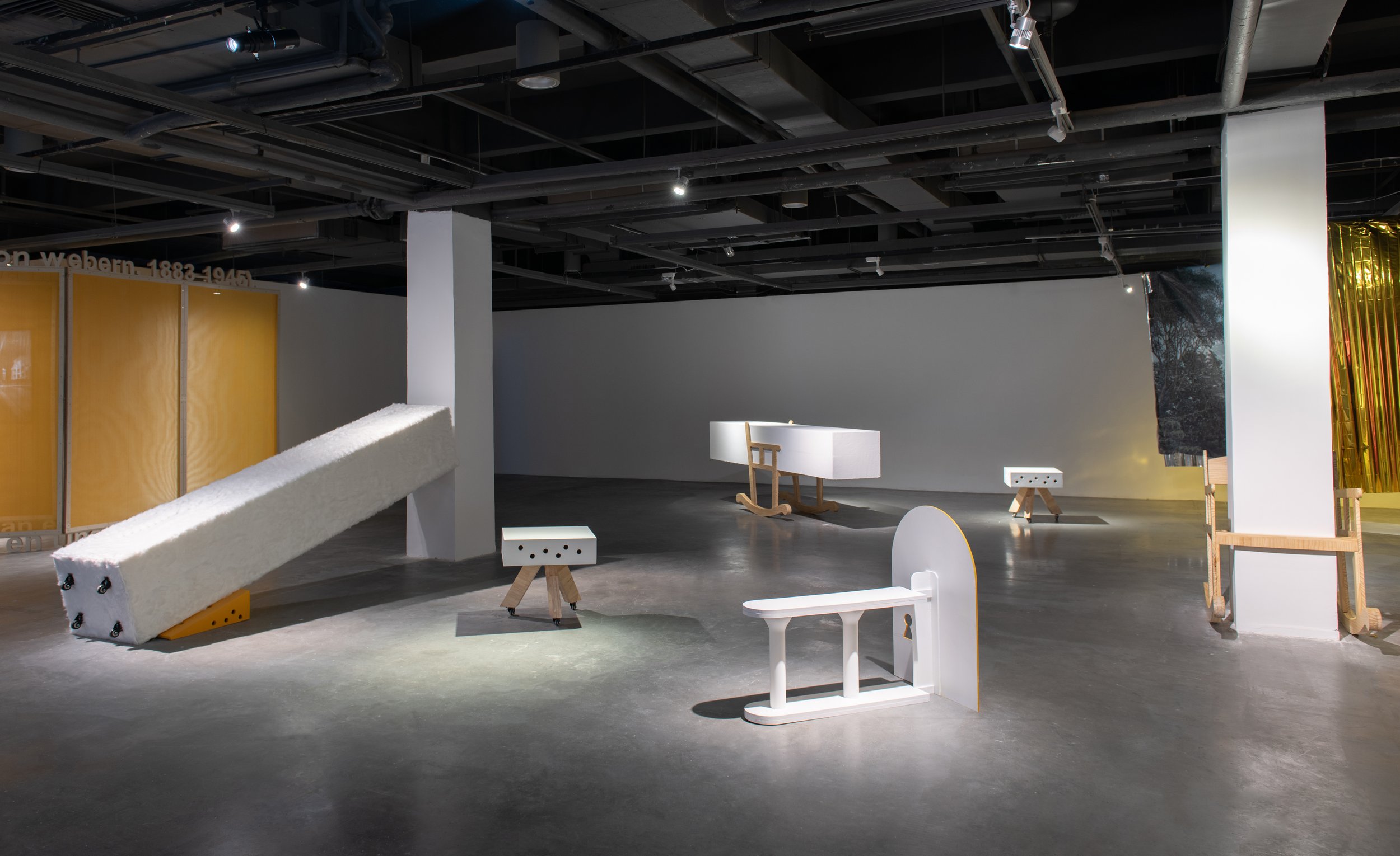 Wide gallery installation in which a large white architectural column tilts diagonally toward the floor among small furniture-like sculptural supports and objects.