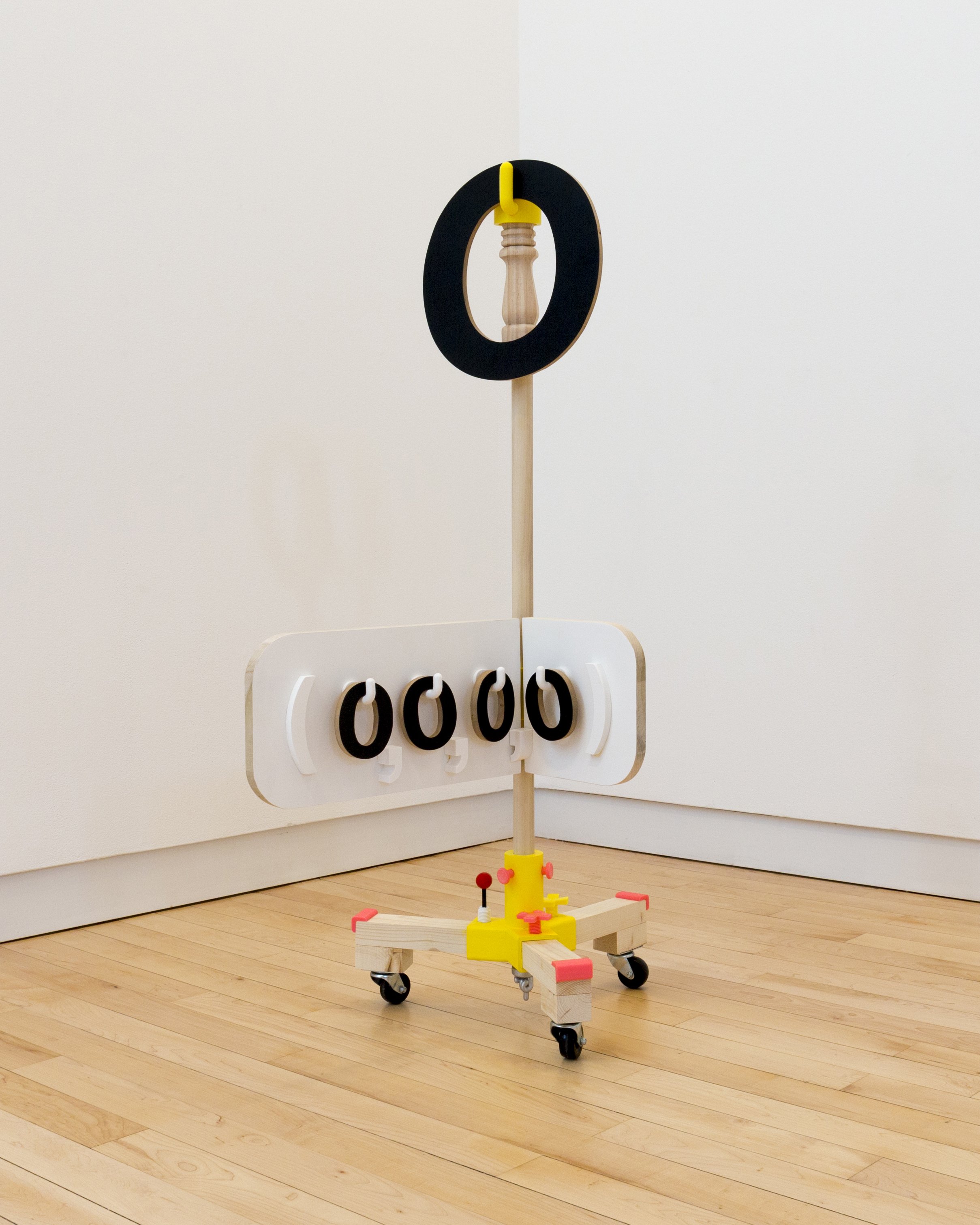 Vertical wheeled sculpture with a black circular ring surrounding a wooden spindle above a white panel displaying four black zero-like shapes.