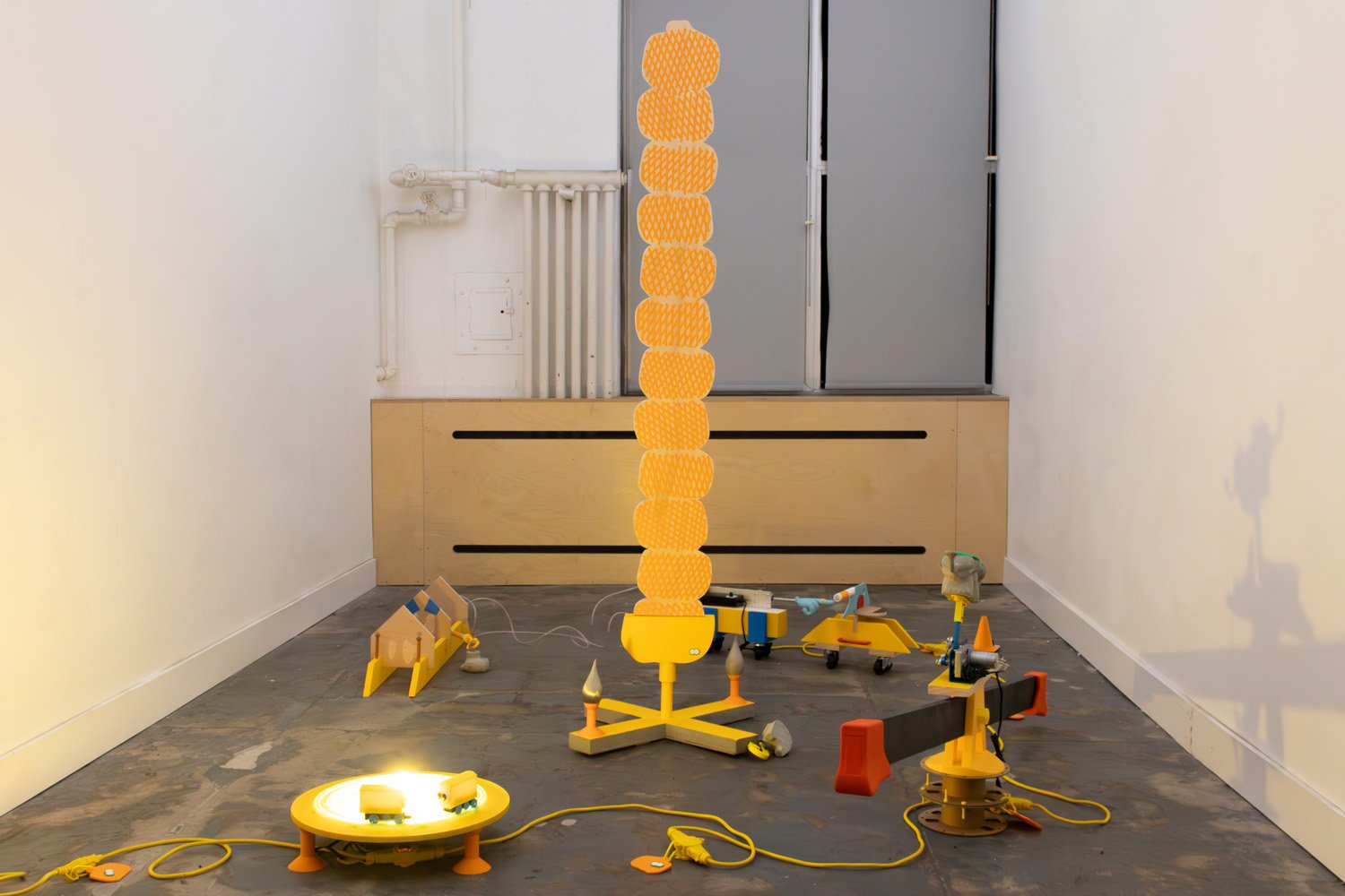 Installation view of brightly colored sculptural machines and tools arranged on a gallery floor around a tall vertical stack of orange forms.