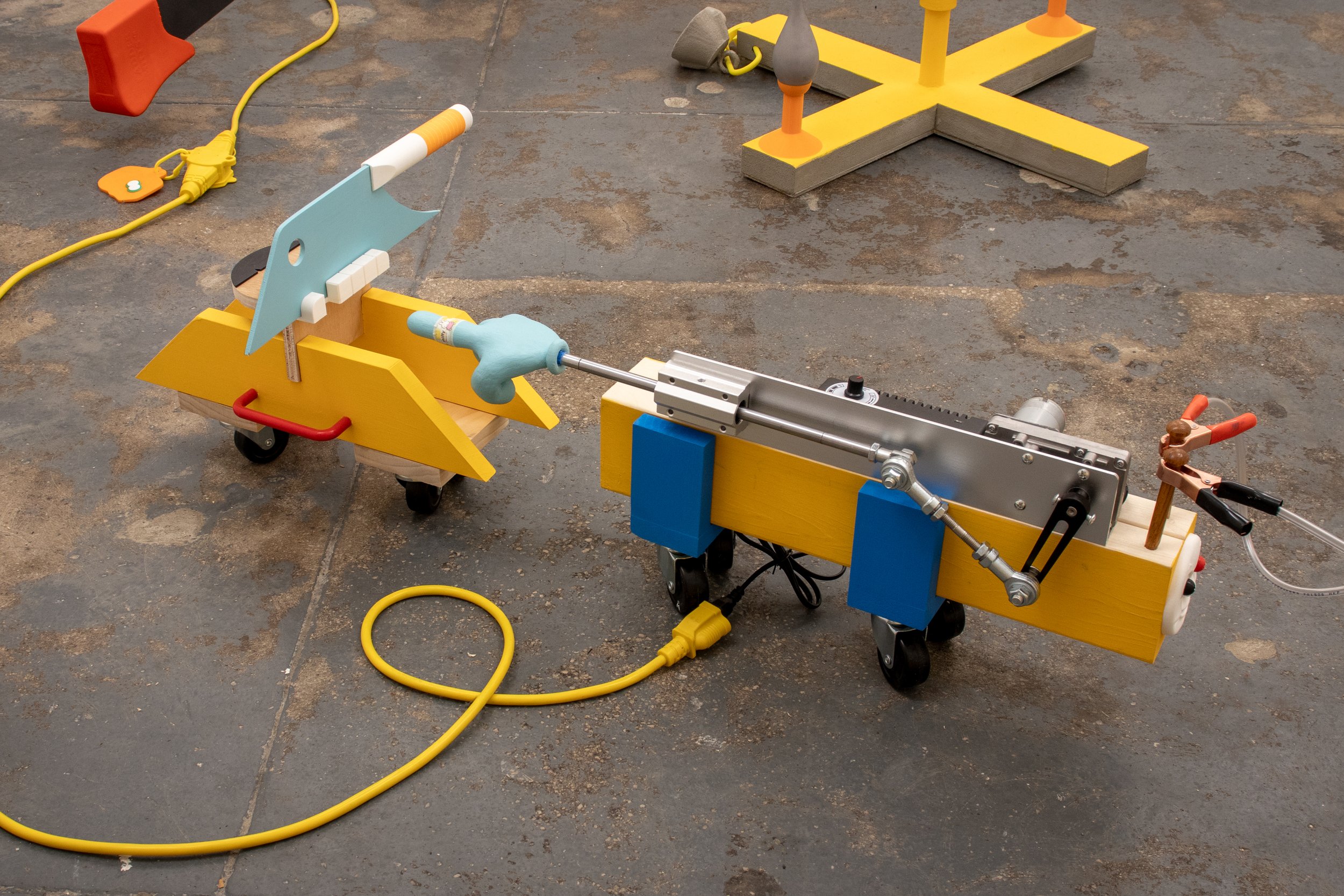 Detail of colorful wheeled sculptural devices made from yellow and blue wood, metal mechanisms, toy-like figures, and electrical cords.