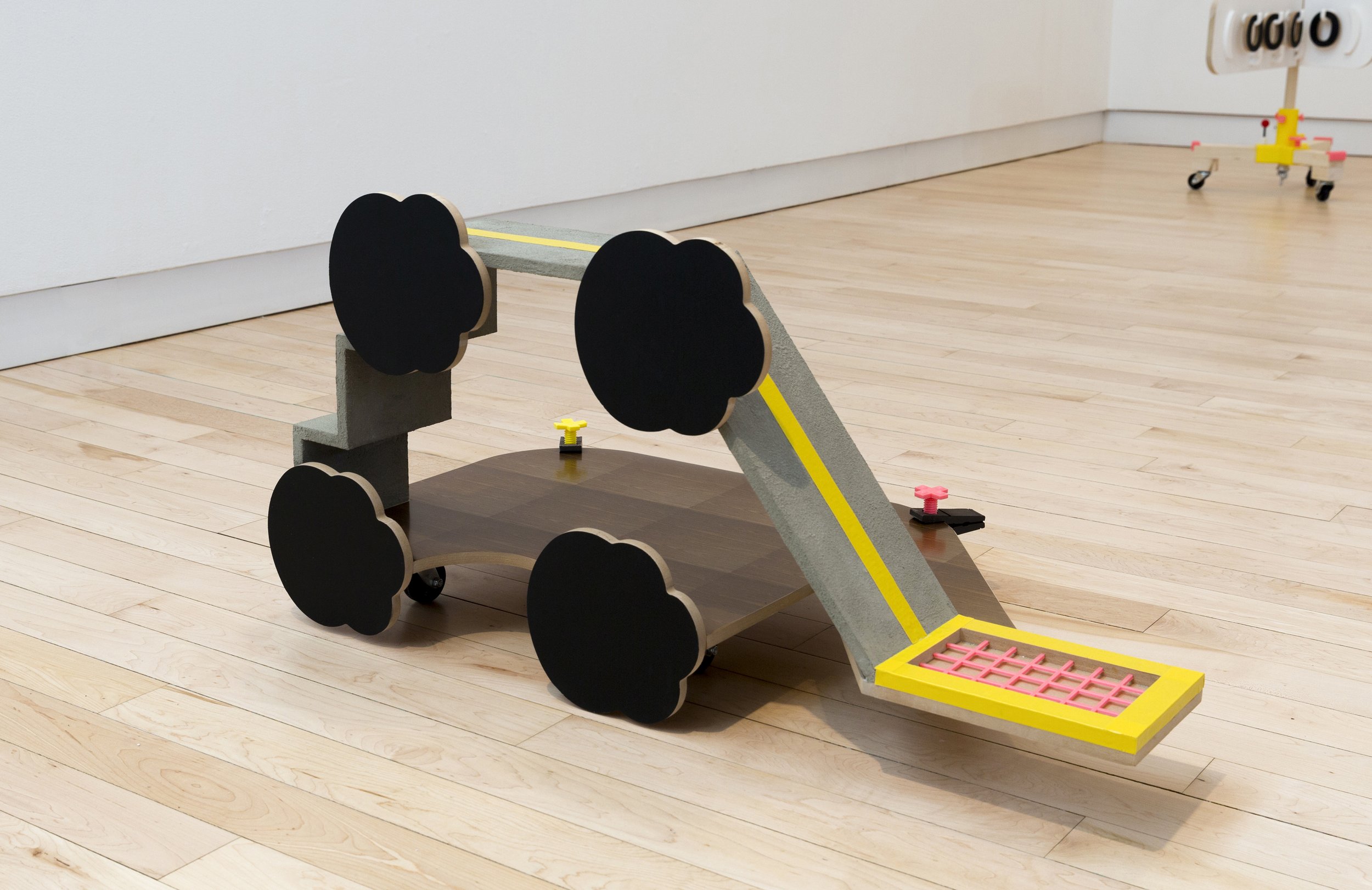 Low wheeled sculpture with four black flower-shaped wheels and a sloping gray ramp edged in bright yellow, resembling a hybrid playground structure and vehicle.