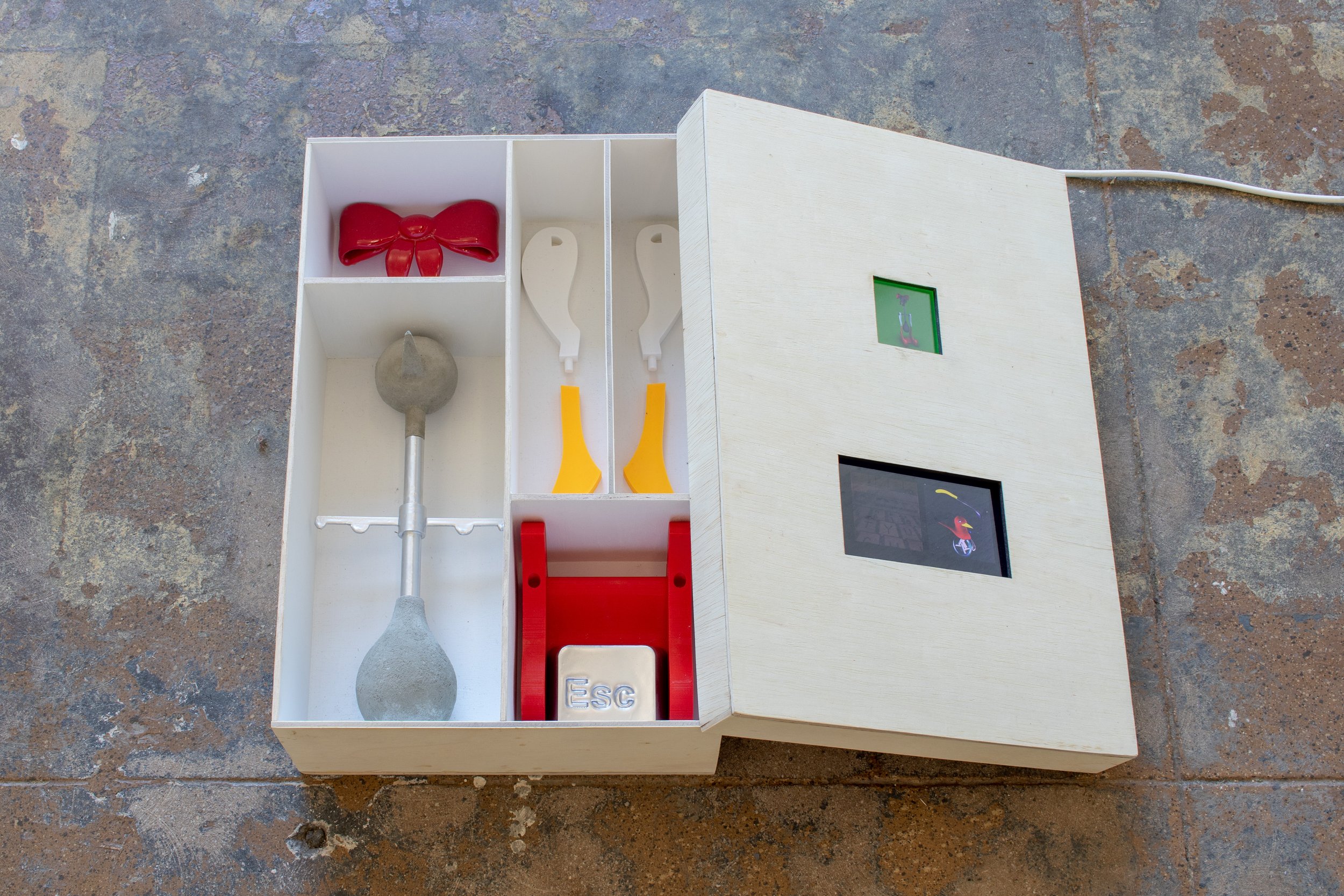 Open wooden model-kit box containing colorful sculptural parts, including a red bow, shovel-like forms, an ESC key, and two small video screens built into the lid.