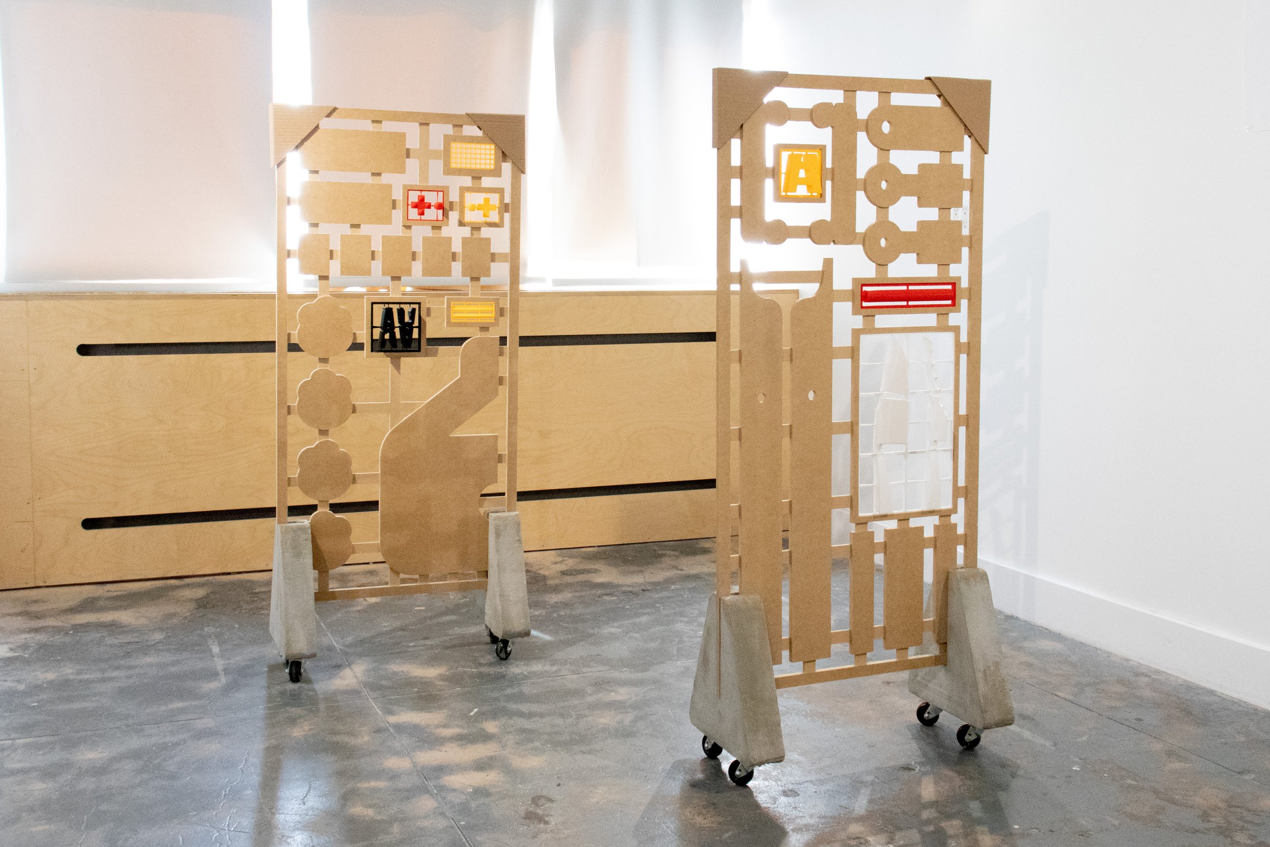 Two tall freestanding model-kit panels on wheeled concrete bases, with cutout component shapes and small colorful plastic parts embedded in the frames.