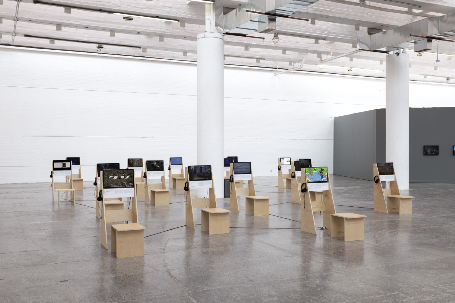Wide installation view of Projected Ecologies showing multiple video stations, monitors, stools, and a gray partition wall.