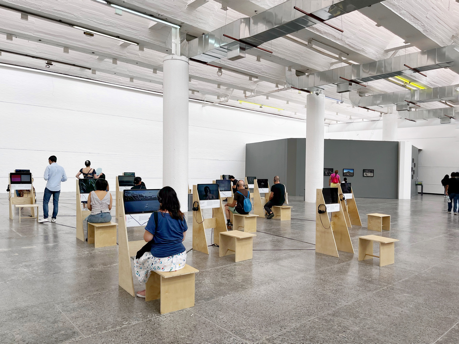 Visitors seated at video stations wearing headphones in the Projected Ecologies exhibition, with a gray partition wall in the background.