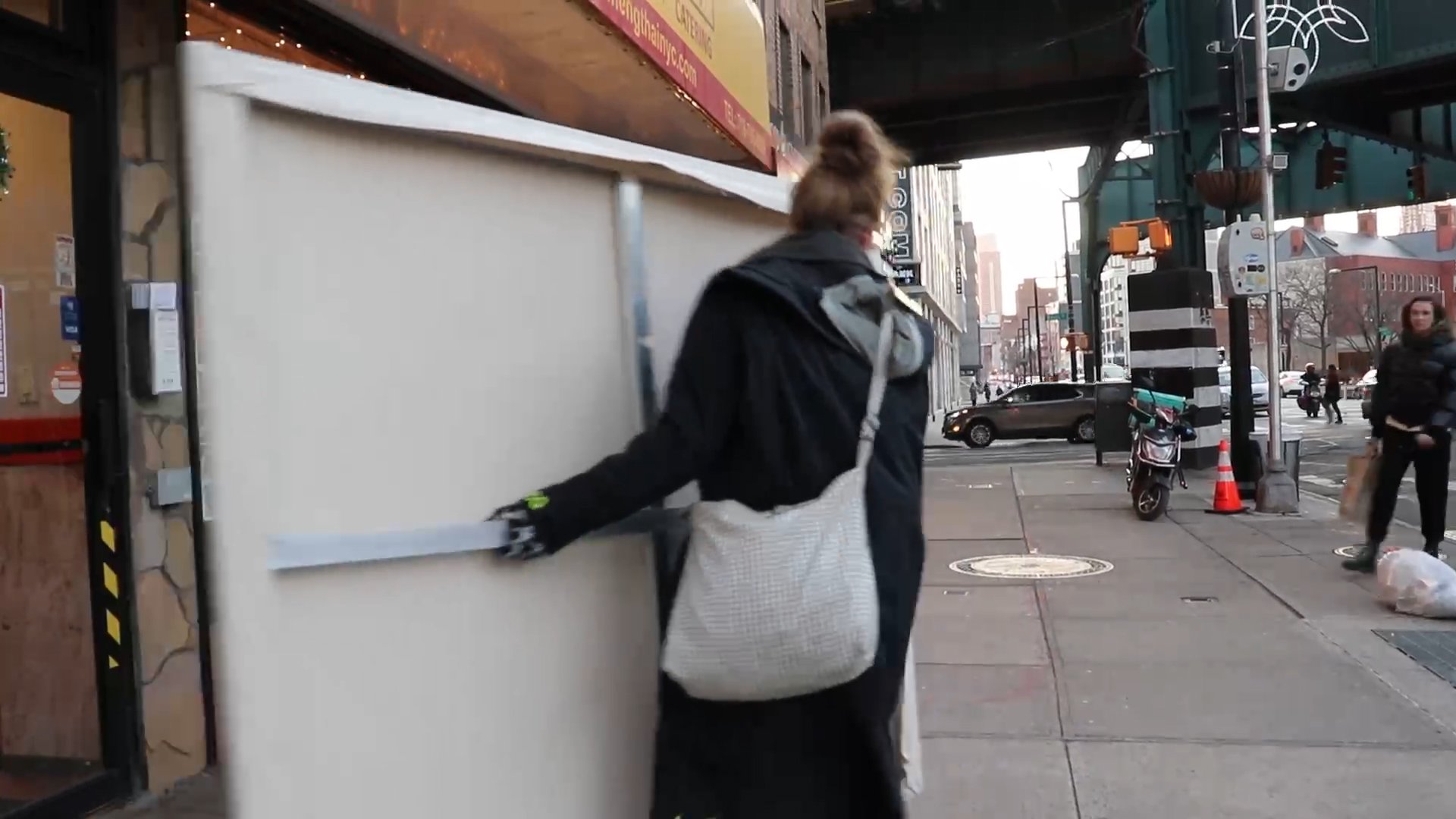 Video still showing a person carrying a large rectangular canvas along a city sidewalk.